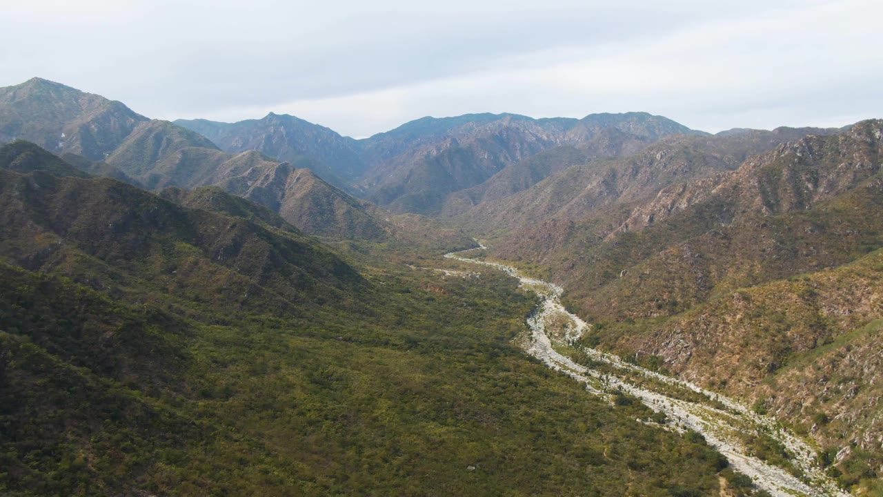 San Dionisio aerial frame showing the riverbed centered lower in the valley, with scrubby green terrain and rocky mountain walls under soft cloud cover, no visible people.