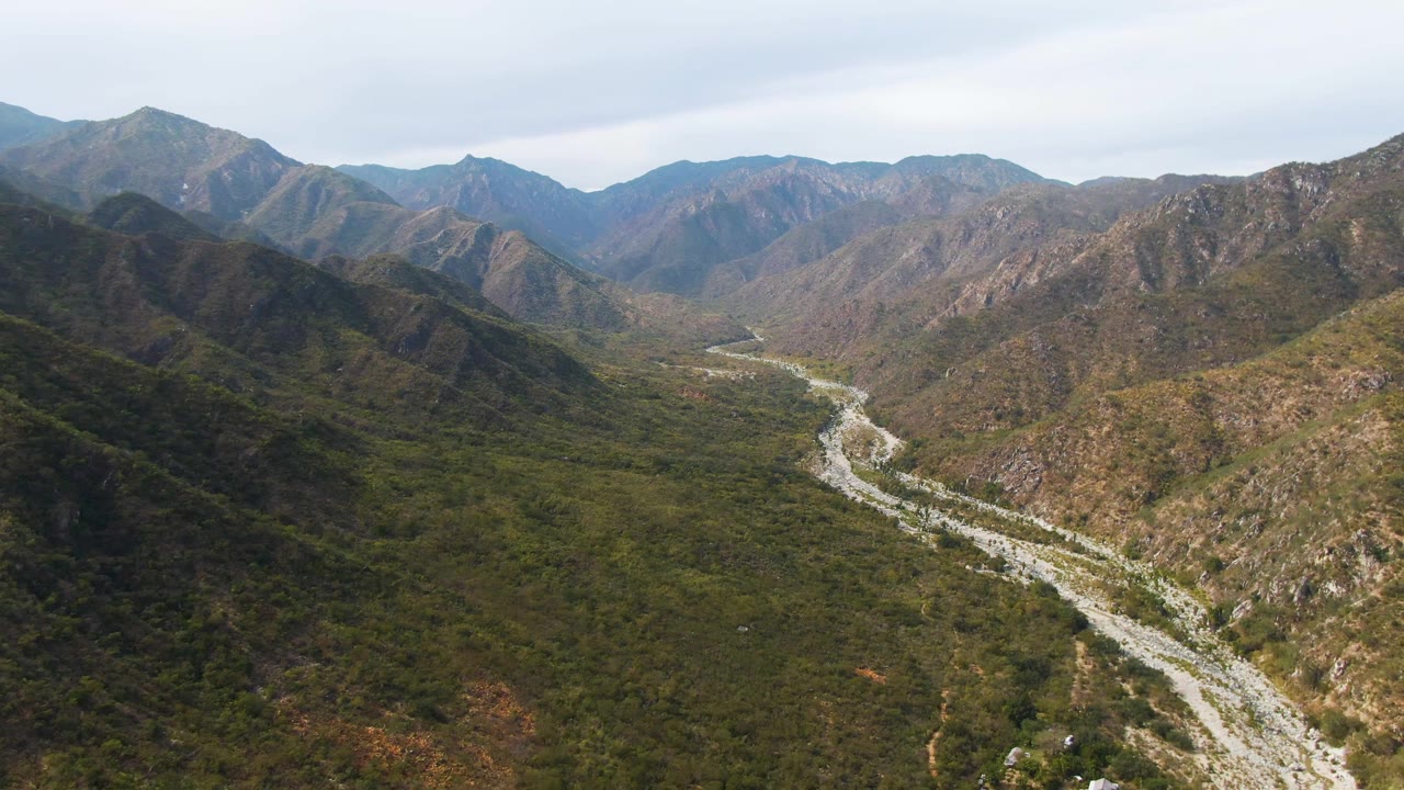 Aerial frame over San Dionisio showing the riverbed crossing the lower valley foreground, with darker green slopes on the left and rocky brown ridges in the distance, no visible people.