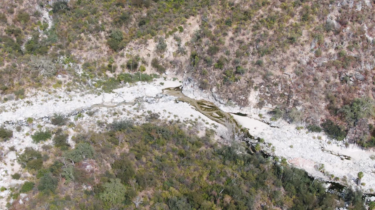 Aerial frame over San Dionisio, Baja California Sur, with dark water visible in a narrow channel beside pale stone and patchy hillside vegetation, no visible people.