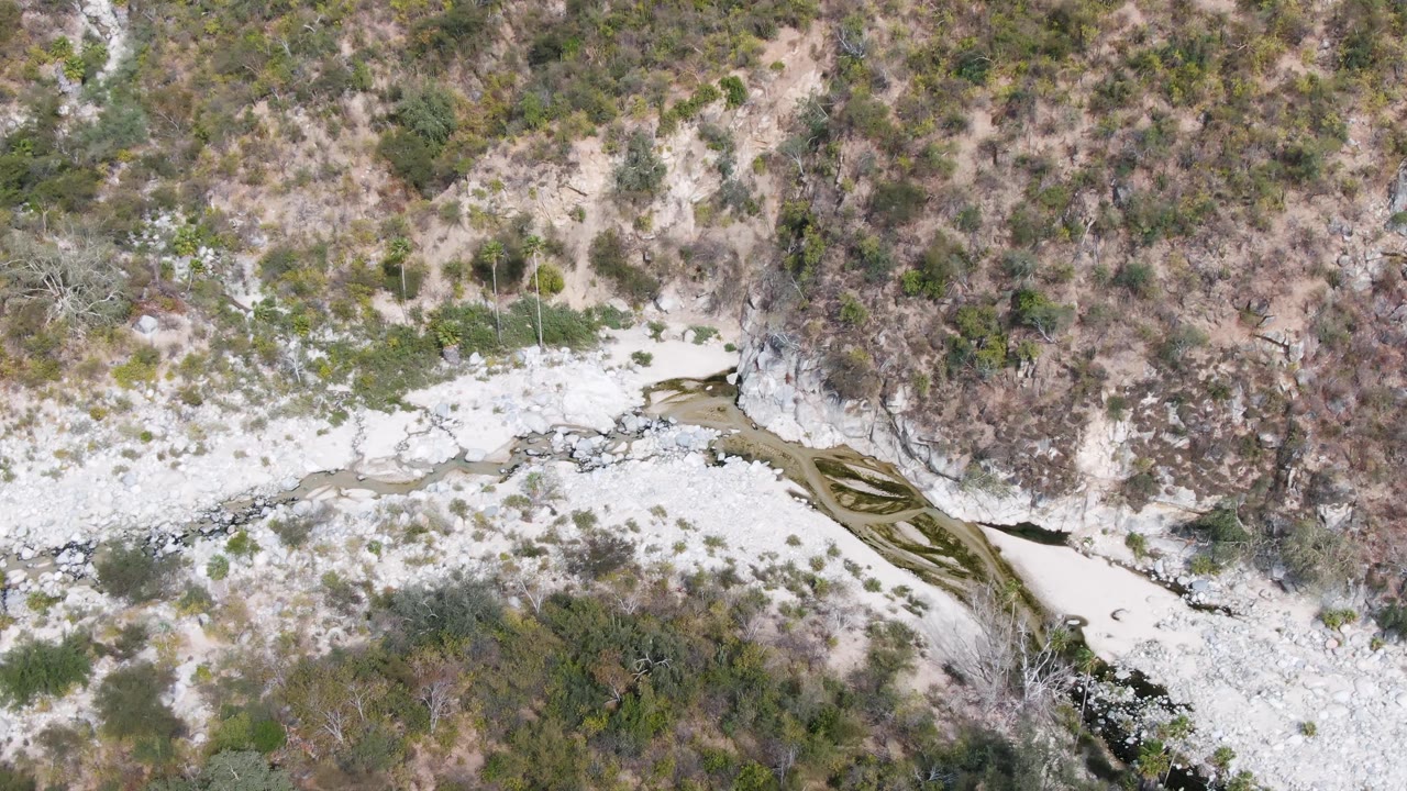 Mid-clip aerial frame in San Dionisio, Baja California Sur showing the dry rocky riverbed, a small dark water flow, scrub patches, and no visible people.