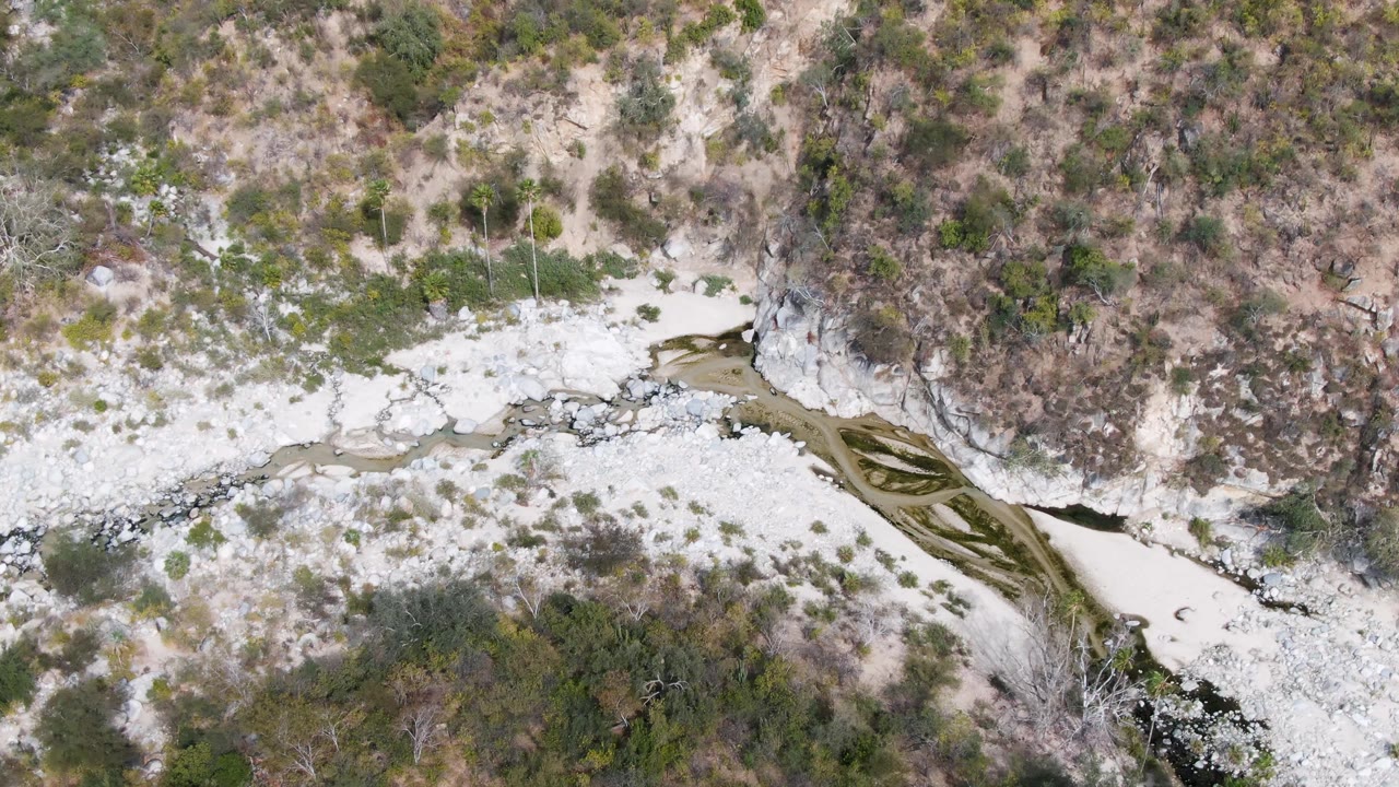 Aerial view of a pale rocky river wash with dark water and sparse scrub.