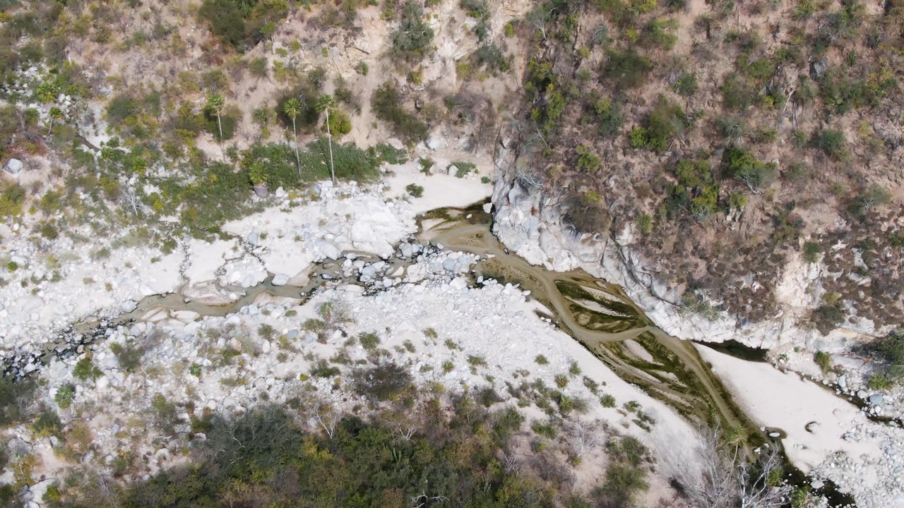 Fourth frame over San Dionisio, Baja California Sur showing the river wash continuing through rocky banks, brown scrub, and scattered green plants with no visible people.