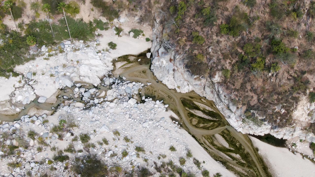 A high overhead view in San Dionisio, Baja California Sur shows brown water curving through a white rock riverbed below a vegetated cliff edge, with no visible people.