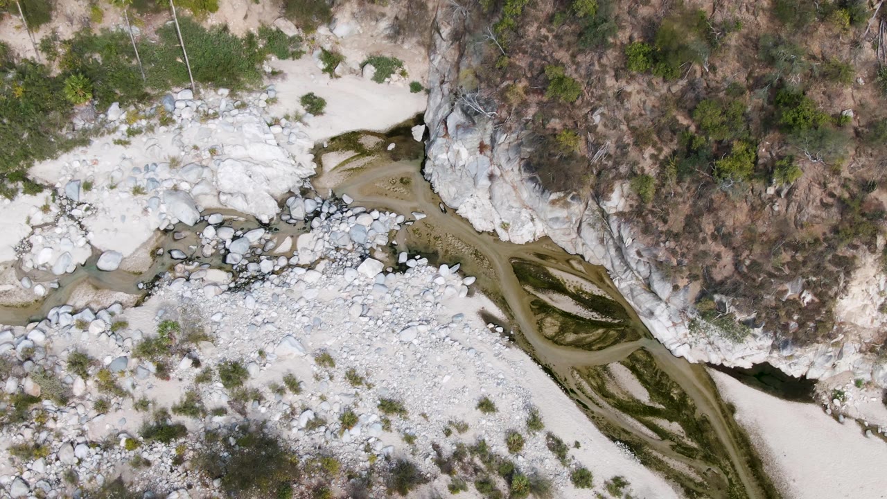 The aerial frame over San Dionisio, Baja California Sur centers the pale stone riverbed and dark brown channel beside the cliff wall, with sparse green vegetation and no visible people.