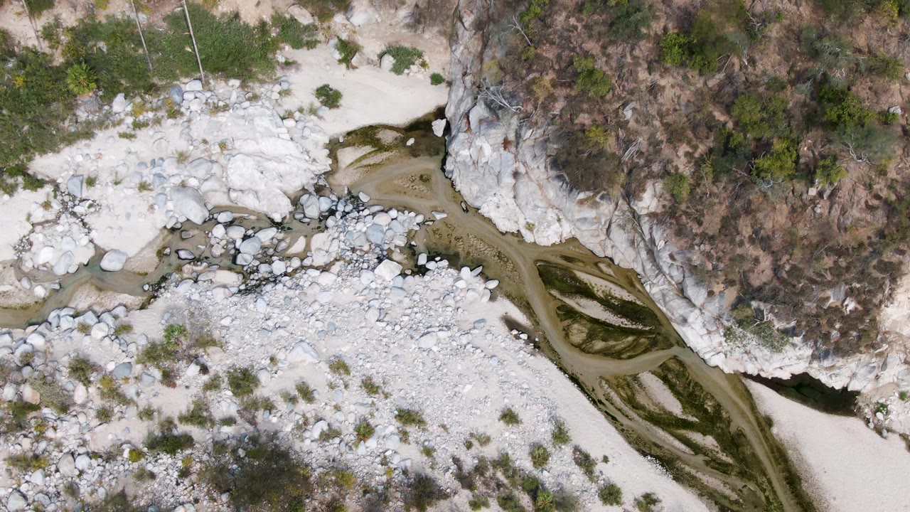 An overhead view in San Dionisio, Baja California Sur shows the stream running along the base of a scrub-covered cliff, with white boulders and sand bars visible and no visible people.
