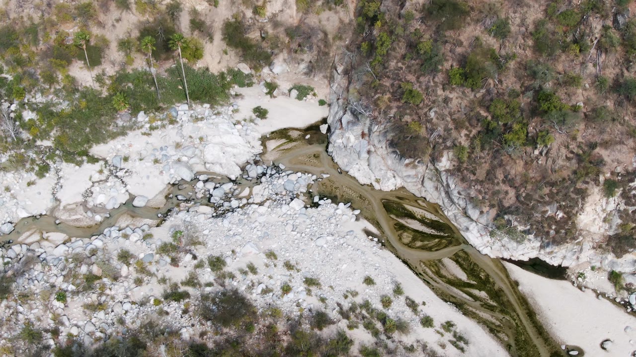 Top-down aerial view in San Dionisio, Baja California Sur showing braided water lanes beside a broad pale rock wash and a dry scrub-covered hillside, with no visible people.