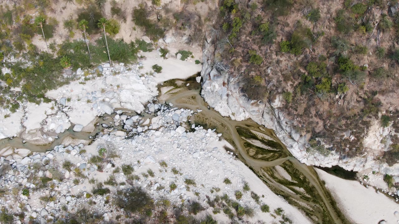 Aerial frame in San Dionisio, Baja California Sur where the diagonal stream runs along boulders and a pale gravel bar below dry brush and rocky terrain, with no visible people.