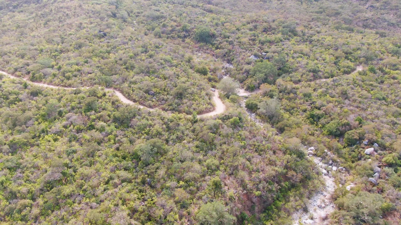 Aerial frame over San Dionisio, Baja California Sur showing the S-shaped dirt road above scrub-covered slopes and a narrow rocky stream to the right, with no visible people.