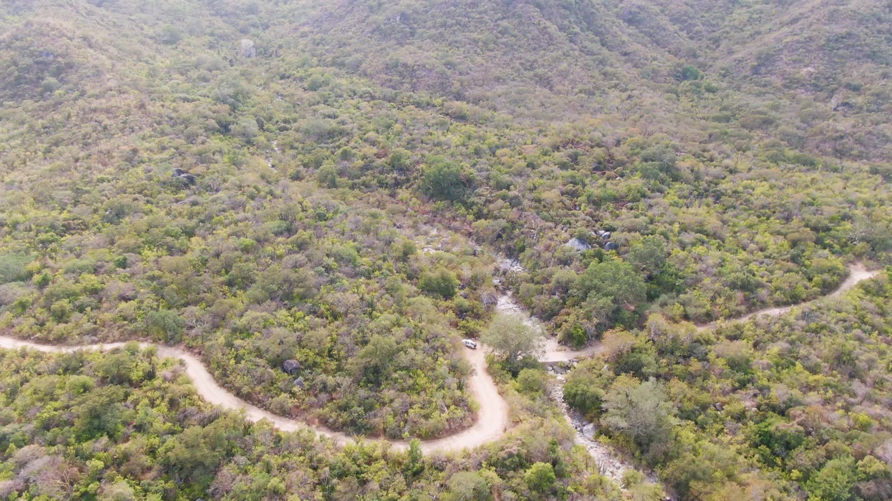 Aerial view in San Dionisio, Baja California Sur, with the road bending across dense hillside vegetation and a narrow rocky wash visible nearby, with no visible people.