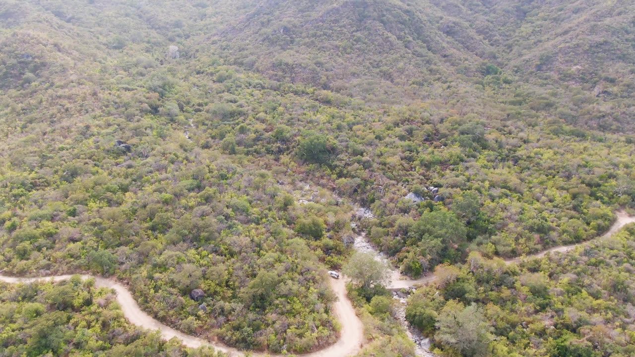 High aerial frame from San Dionisio, Baja California Sur, showing a pale track winding below rounded green and brown hills with scattered rocks and no visible people.