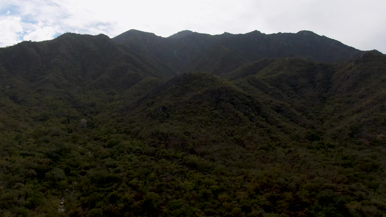 Wide aerial view of dark forested mountain ridges under cloudy sky.