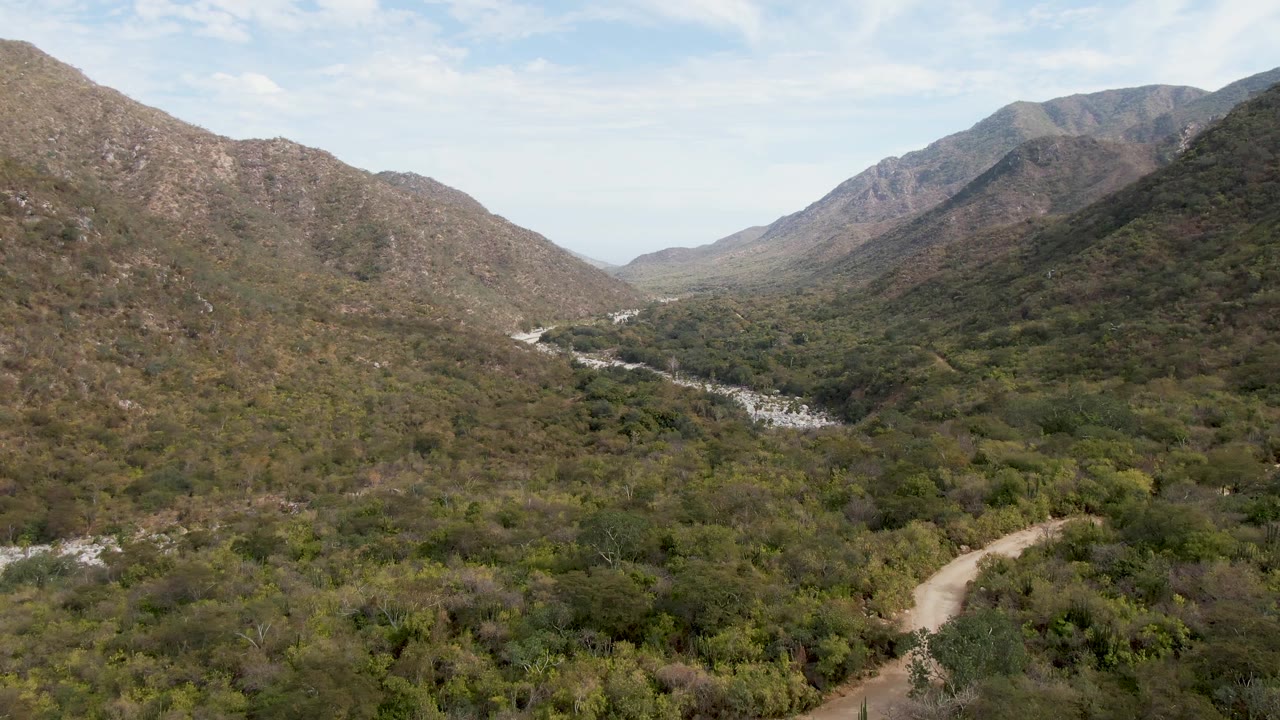 Opening aerial frame over San Dionisio, Baja California Sur, showing rocky hills around a green valley, a pale riverbed at left, a dirt road at lower right, and no visible people.
