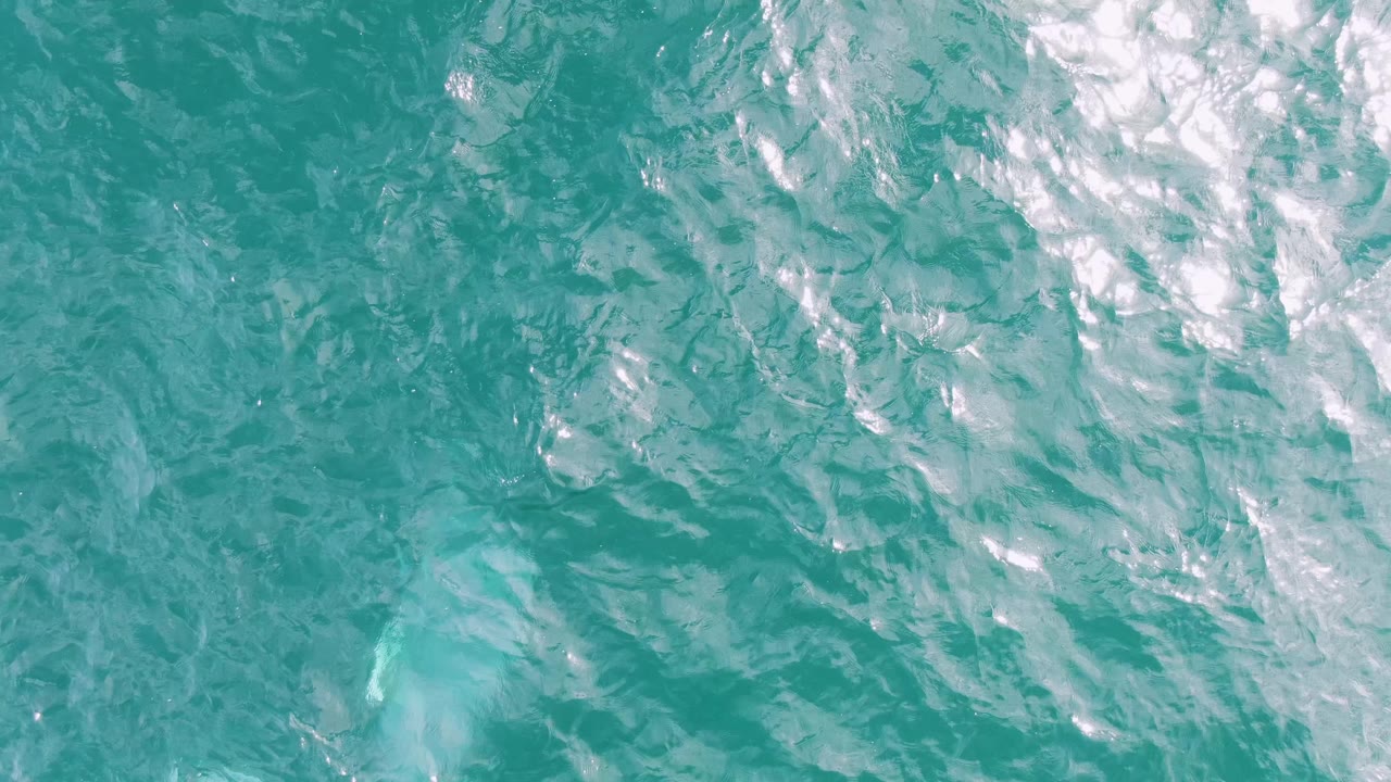 Overhead view of turquoise ocean water at Shipwrecks East Cape, Baja California Sur, with a faint pale blue trail under rippled reflections and no visible people.