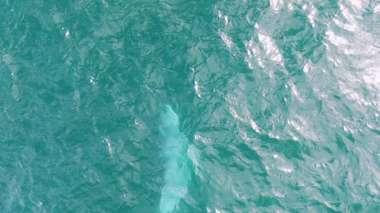 Aerial-looking view of turquoise water at Shipwrecks East Cape, Baja California Sur, with textured ripples and a pale blue trail sitting lower in the frame, with no visible people.