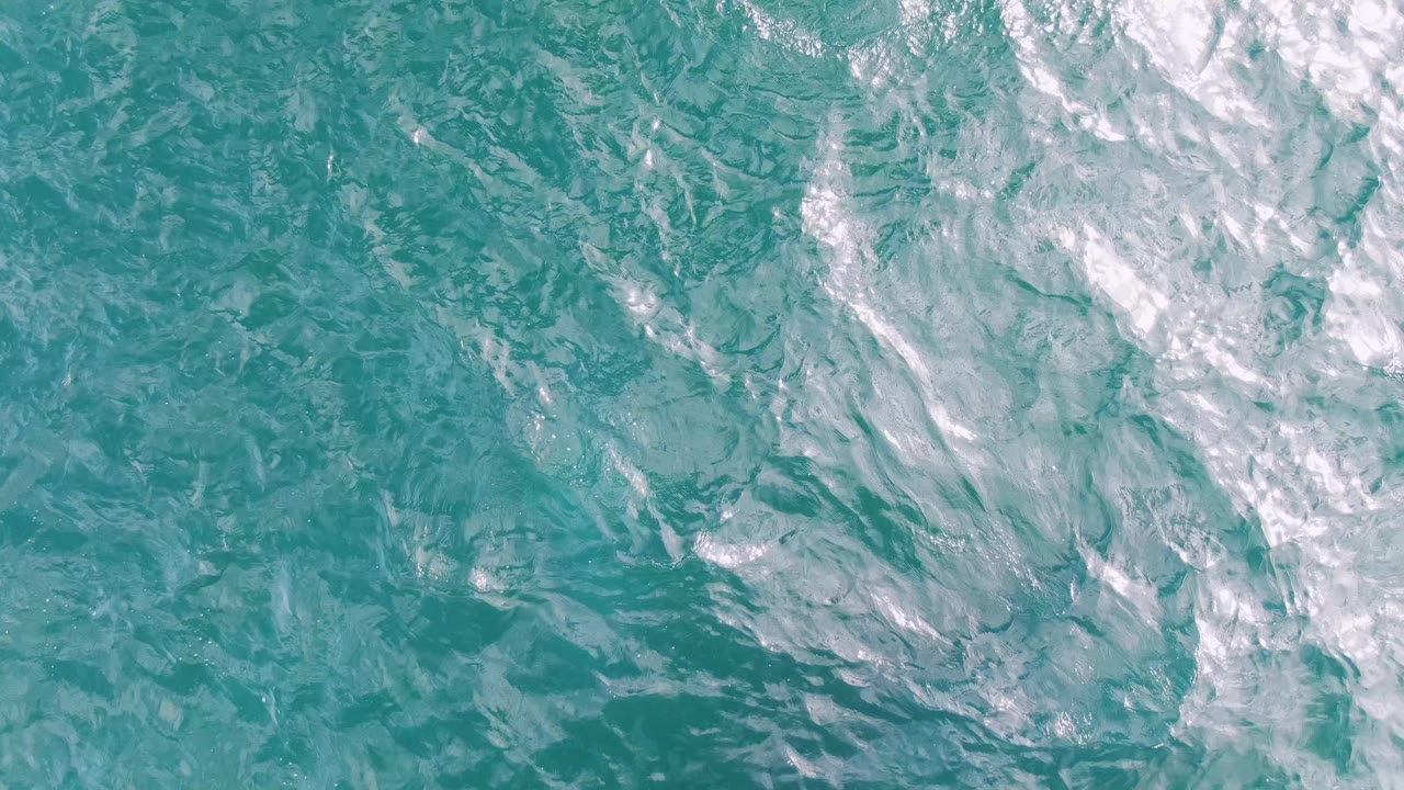 Overhead aerial frame of clear turquoise water at Shipwrecks, Baja California Sur, with foamy white wake streaks crossing the upper right area and no visible people.