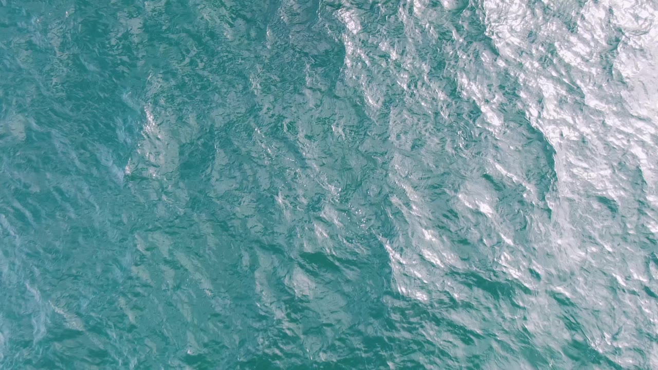 Overhead ocean surface at Shipwrecks, Baja California Sur, with teal water texture and bright white foam concentrated along the right edge, with no visible people.