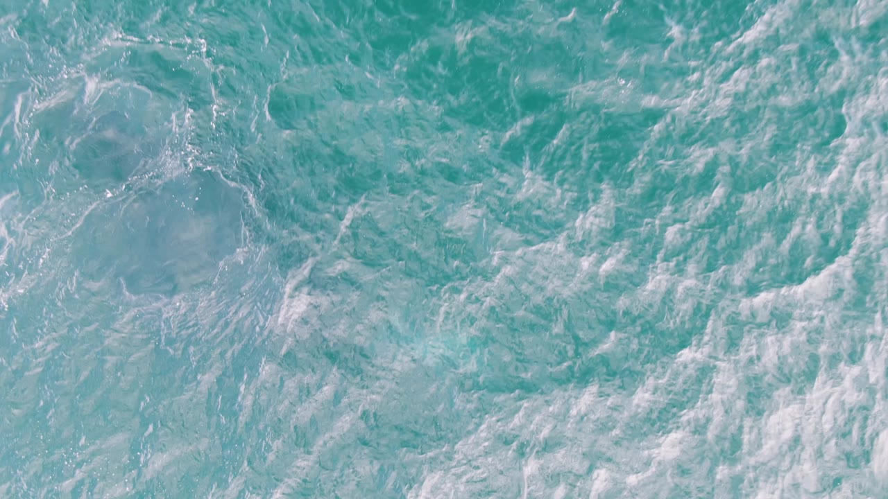 Overhead sea surface at Shipwrecks, Baja California Sur, where broader white foam patches drift across turquoise water with no visible people.
