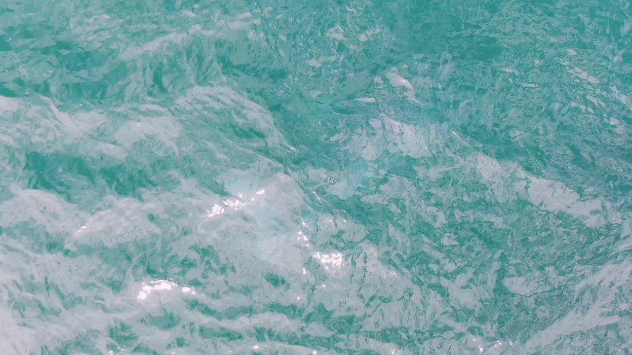 Clear blue-green ocean surface at Shipwrecks, Baja California Sur, with broken white foam patches and small light glints, no visible people.