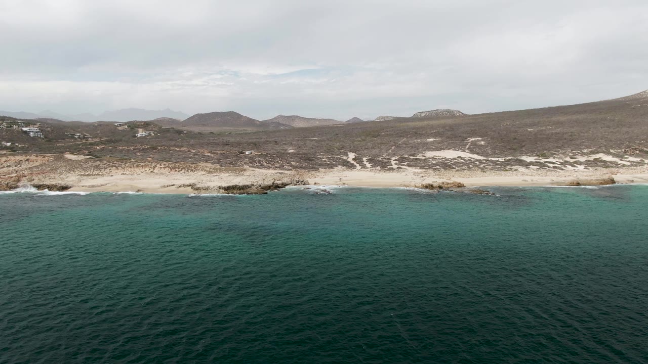 Opening aerial frame over Shipwrecks, Baja California Sur, with deep teal water in front, pale surf along a mixed sand and rock shoreline, dry hills beyond, and no visible people.