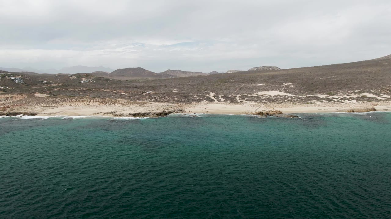 Mid-clip aerial view of Shipwrecks, Baja California Sur, where small waves break over a sandy and rocky shoreline below scrubby hills, with no visible people.