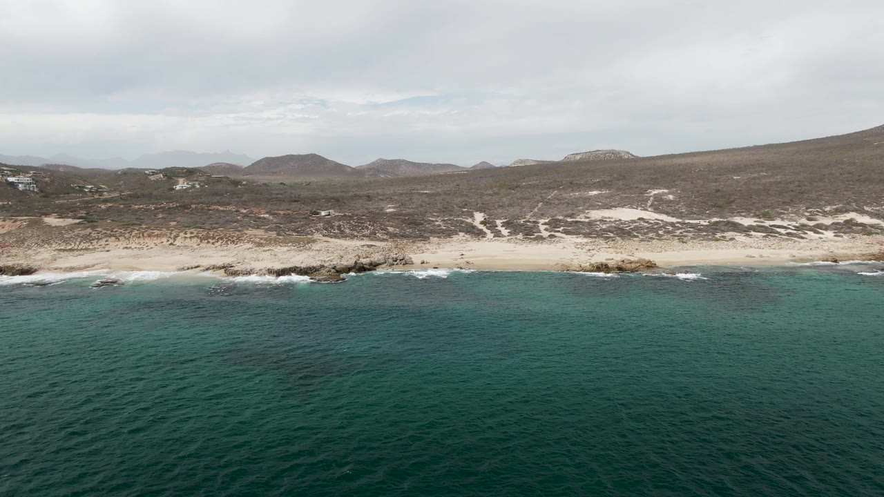 Wide aerial frame over Shipwrecks, Baja California Sur, Mexico, with turquoise water in the foreground, pale beach and dark rocks at shore, low hills beyond, and no visible people.