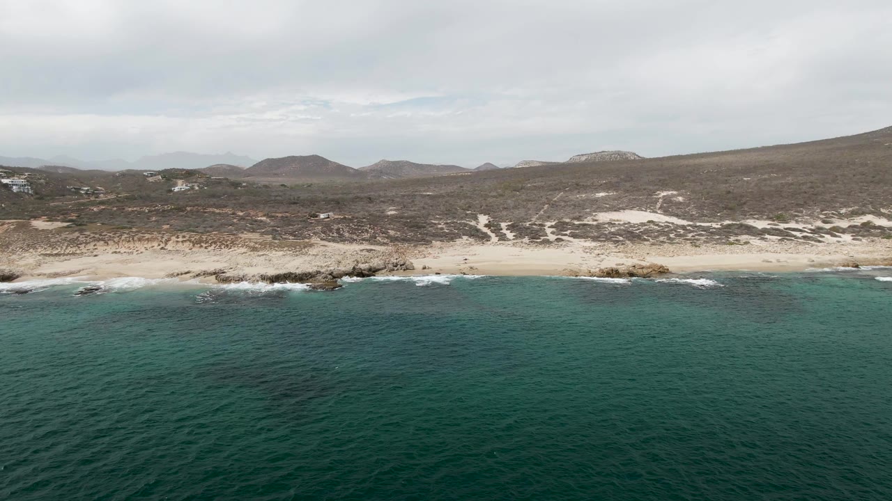 Aerial shoreline view at Shipwrecks in Baja California Sur, Mexico, with the surf line shifted along the sandy beach, rocky points near the water, distant hills, cloudy sky, and no visible people.