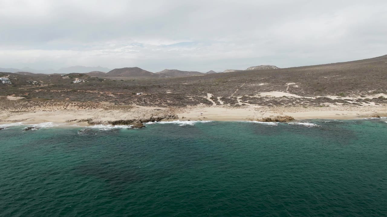 Mid-clip aerial view of Shipwrecks, Baja California Sur, Mexico, showing green-blue ocean across the foreground, a pale beach with scattered rocks, low brown hills, overcast clouds, and no visible people.