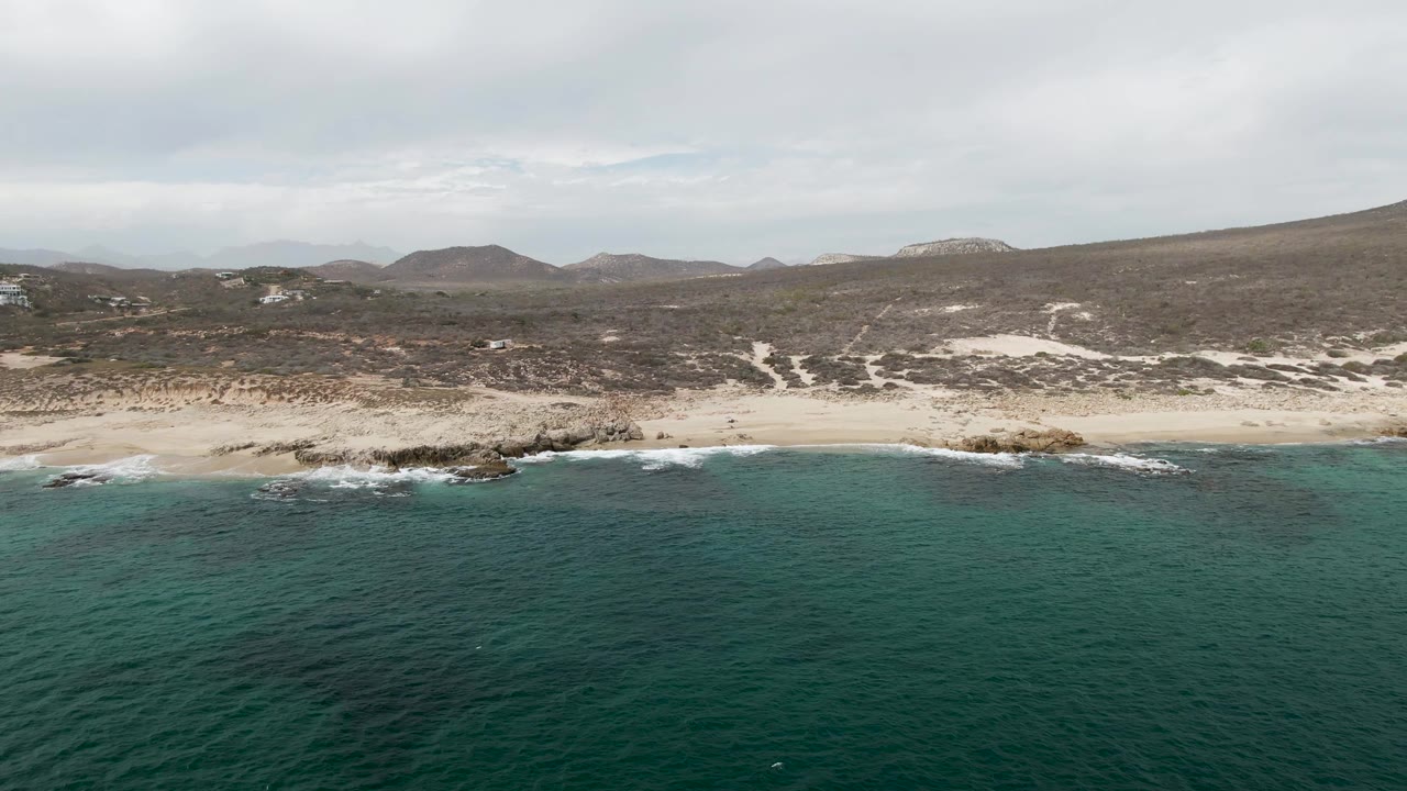 Wide drone view of a rocky sandy coast with turquoise ocean and low hills.