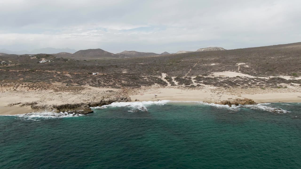 Wide aerial frame of Shipwrecks East Cape BCS Mexico with turquoise ocean in the foreground, pale sand and rocky outcrops along shore, and dry hills under a gray sky.