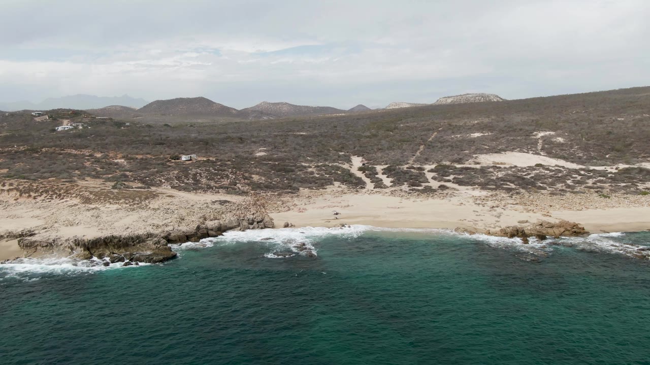 Aerial coastline with waves breaking near rocks and a sandy beach behind.