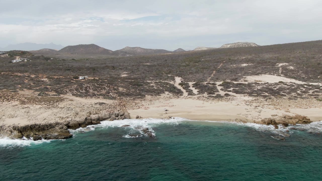 Shipwrecks East Cape BCS Mexico coastline from above, with a rocky point interrupting the sand, turquoise water below, and dry hills and scrub stretching inland.