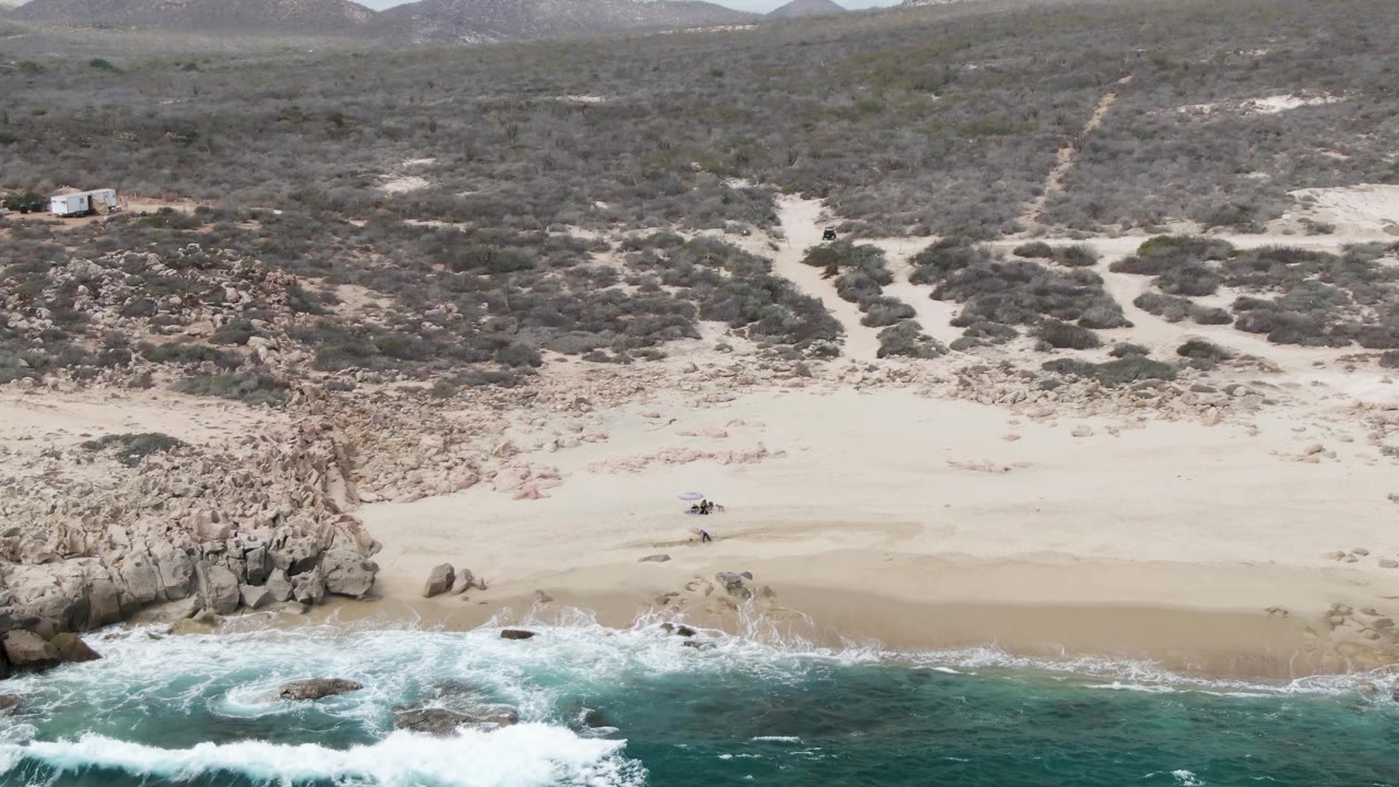 Drone view at Shipwrecks, Baja California Sur with waves curling around shoreline rocks, pale sand above the waterline, scrubland behind the beach, and no visible people.