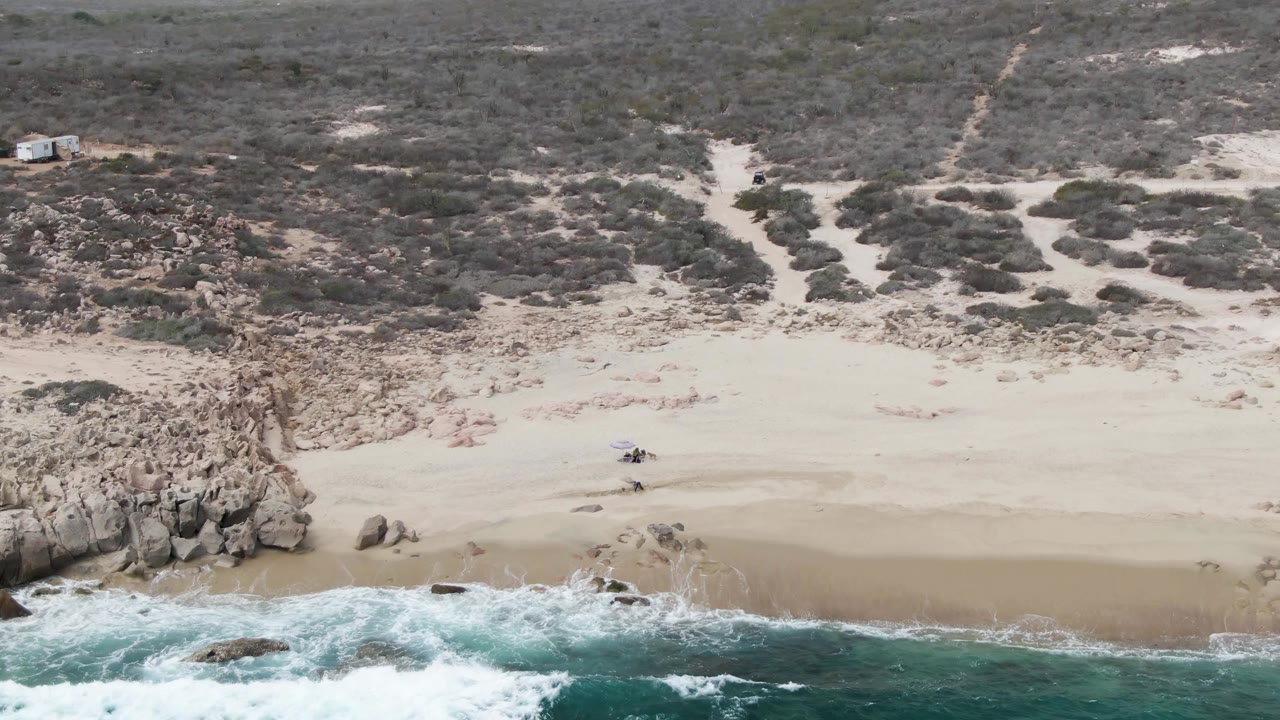 Aerial coastline at Shipwrecks, Baja California Sur showing foamy water across the lower frame, a broad sandy beach, scattered rocks, scrub-covered terrain, and no visible people.