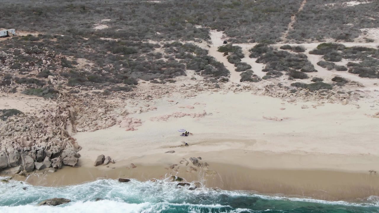 Closer aerial view of sandy beach with rocks, surf, and scrub beyond.