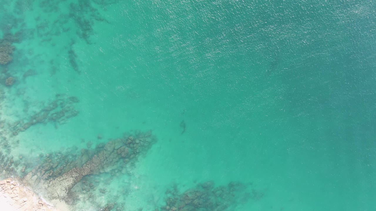 Clear green-blue water with darker submerged rocks and sand along the lower left.