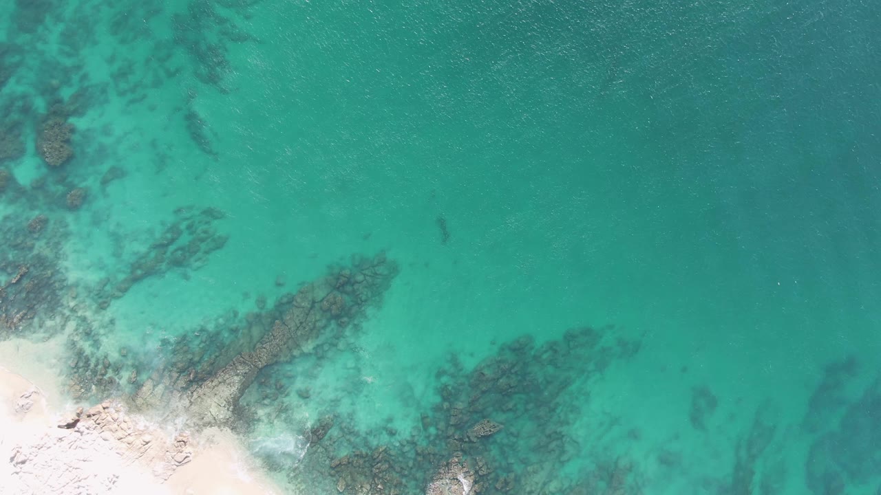 Overhead Buzzards Beach view with white sand brushing the lower left edge, rippled turquoise water across the frame, dark rock patches below, and no visible people.