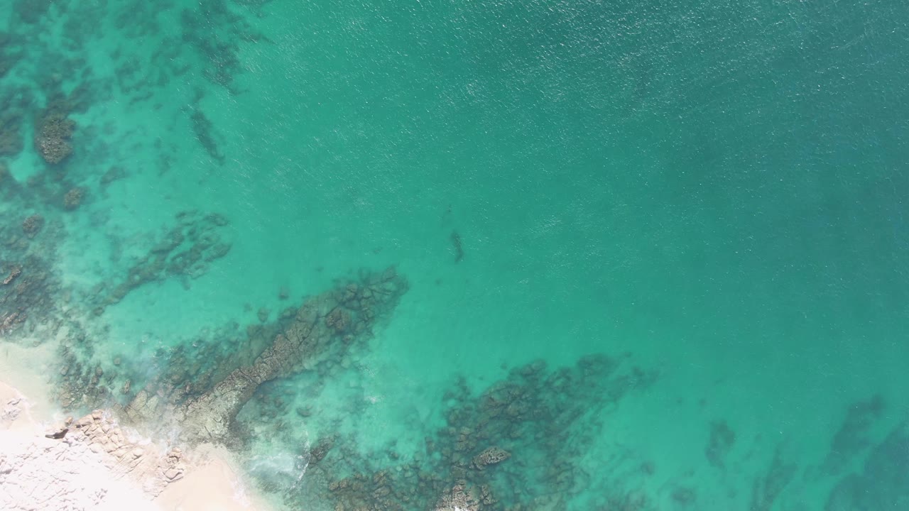 Buzzards Beach aerial still with the sand partly cropped at lower left, broad turquoise shallows across the frame, dark submerged rocks, and no visible people.