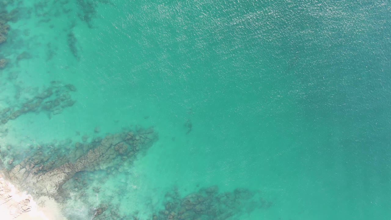 Overhead view at Buzzards Beach, Baja California Sur, showing turquoise water over dark submerged rocks with pale sand entering from the lower left and no visible people.