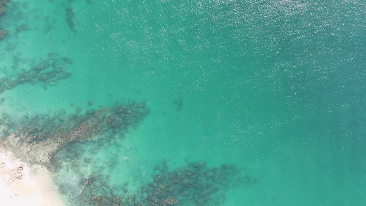 High overhead view at Buzzards Beach, Baja California Sur, with sandy shore cropped at the lower left, darker underwater rocks crossing the shallows, and no visible people.