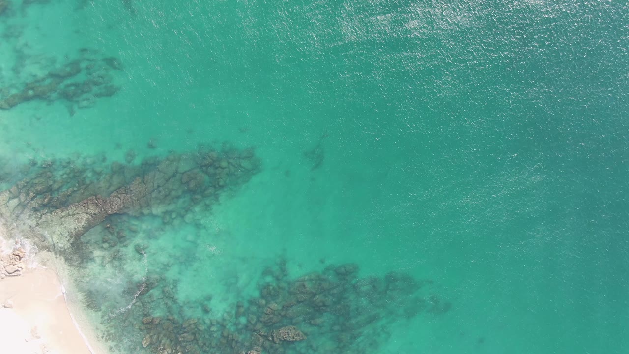Turquoise ocean over submerged rocks beside a pale sandy beach edge.