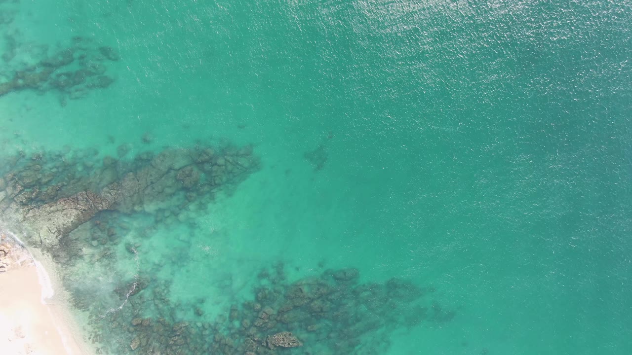 At Buzzards Beach in Baja California Sur, bright turquoise water spreads across most of the frame with dark submerged rocks and a small sand corner, with no visible people.