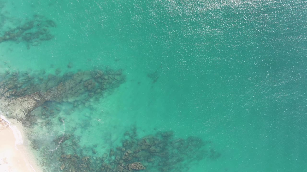 At 0:03, an aerial view at Buzzards Beach shows pale sand along the lower left and dark underwater rock bands beneath turquoise water, with no visible people.