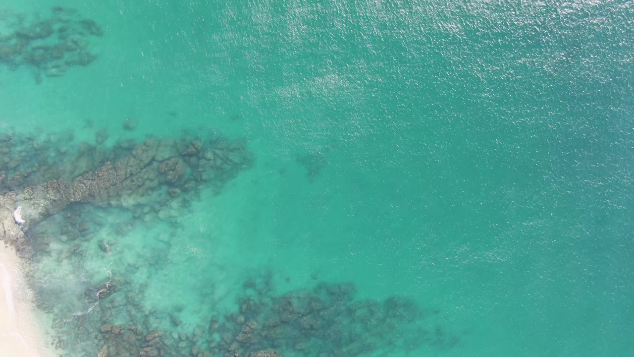 Second frame from Buzzards Beach in Baja California Sur with the sandy shore still visible at lower left, dark underwater rocks crossing the water, and no visible people.