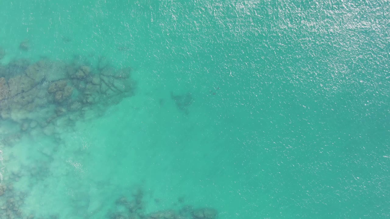 Aerial frame from Buzzards Beach showing a darker submerged rock formation near the upper center beneath rippled turquoise water, with no visible people.