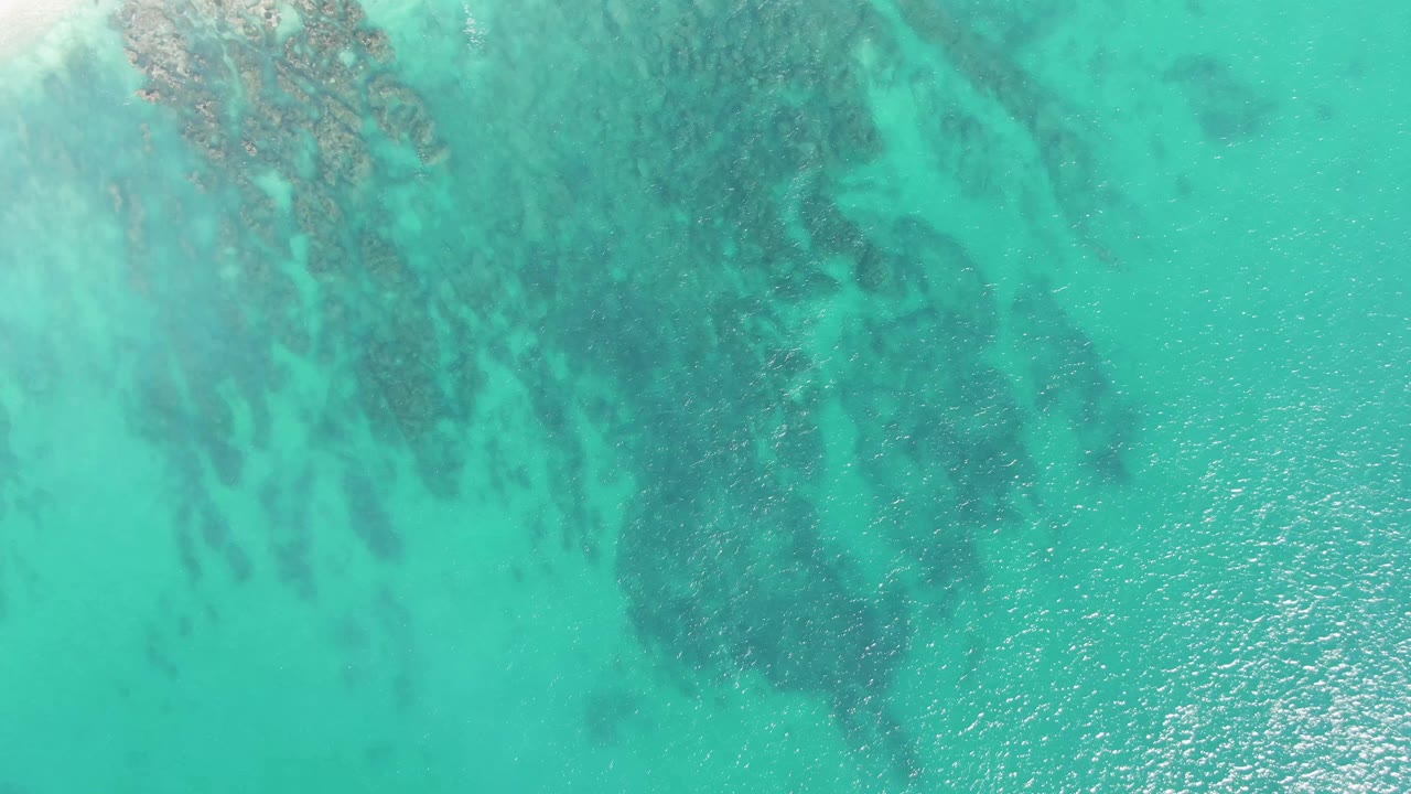 Turquoise shallow ocean with darker submerged rocks and sunlit ripples.