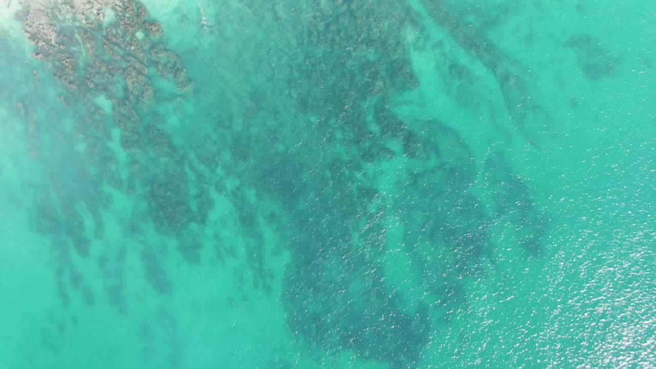 Aerial frame over Buzzards Beach water showing broad turquoise shallows, dark seabed formations, scattered surface sparkle, and no visible people.