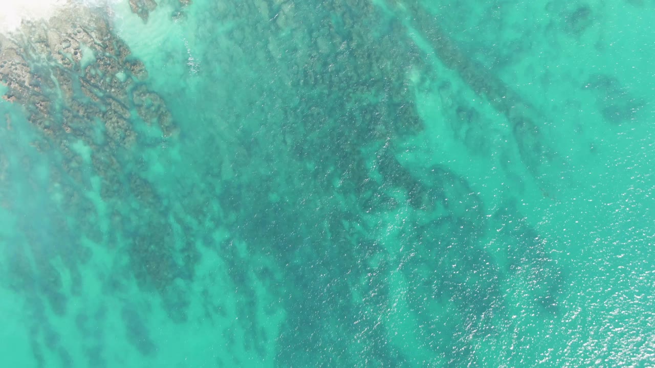 Top-down Buzzards Beach frame with turquoise water, darker underwater rock bands running through the shallows, bright surface texture, and no visible people.