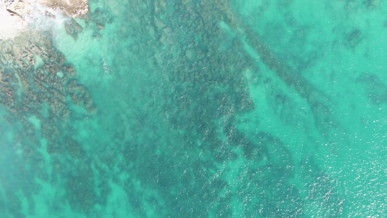 Aerial view at Buzzards Beach where a pale sandy rocky shoreline begins to enter from the left beside clear turquoise water, with no visible people.