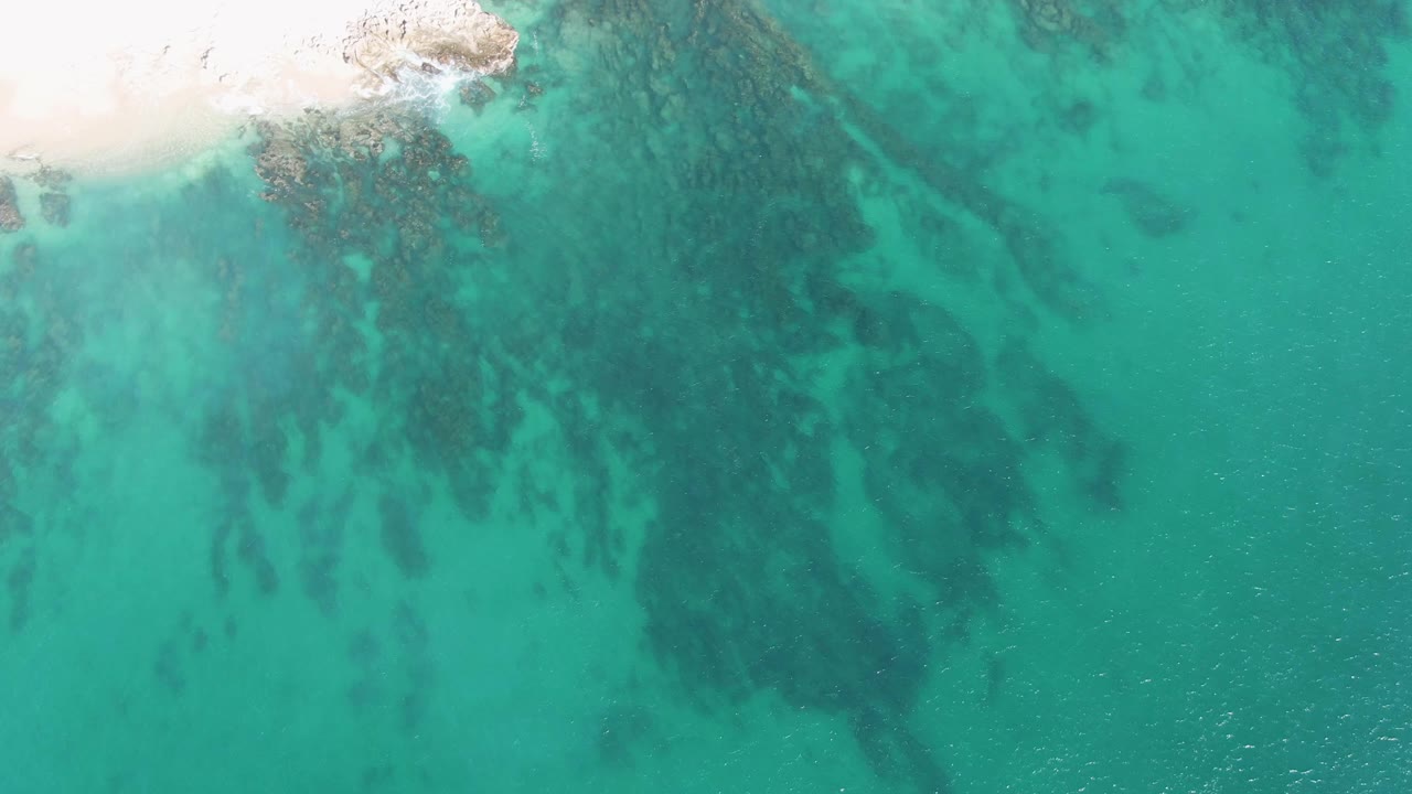 Overhead view at Buzzards Beach with the sandy beach along the top edge, mottled submerged rock patches below the water, and no visible people.