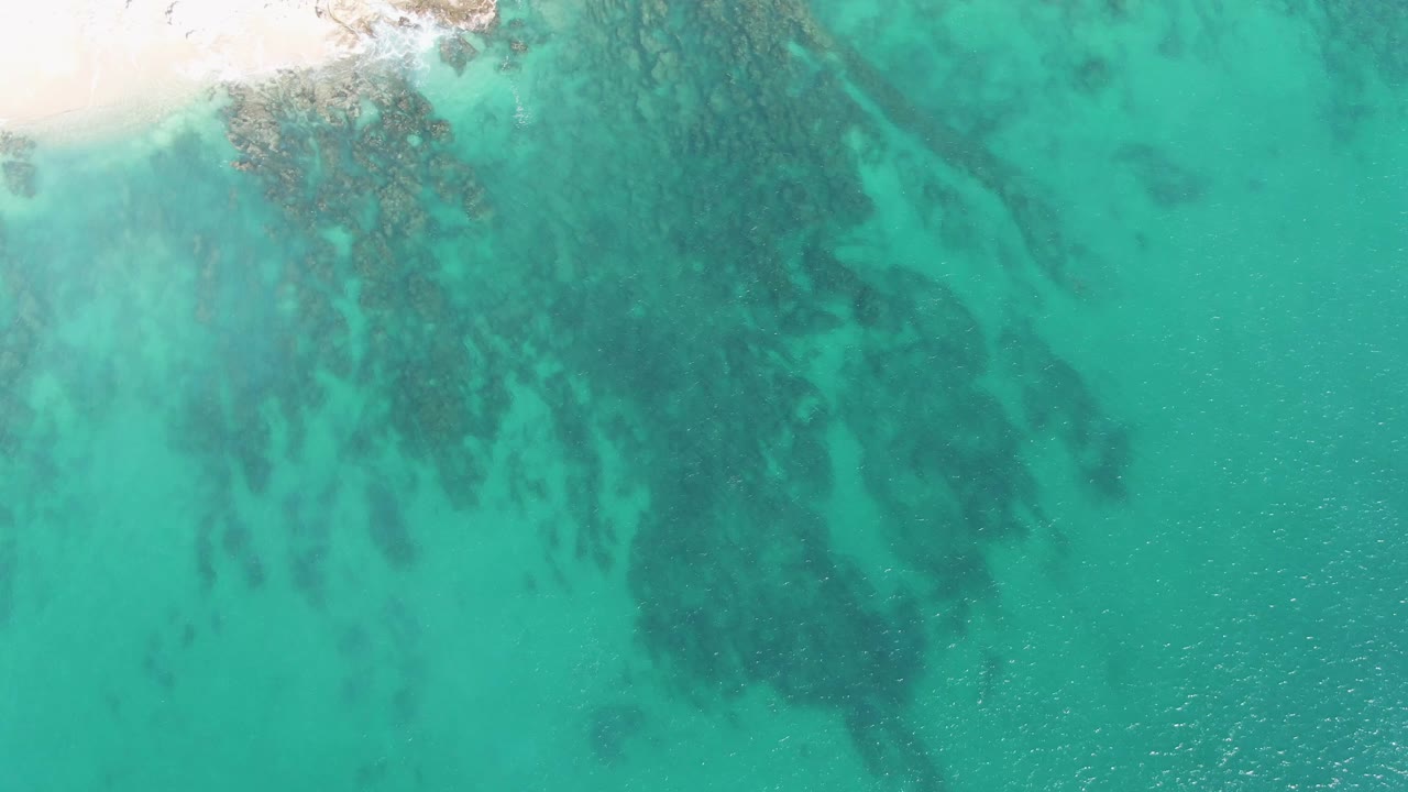 Buzzards Beach aerial frame with a small corner of pale sand, broad turquoise water, darker submerged rocky textures, and no visible people.